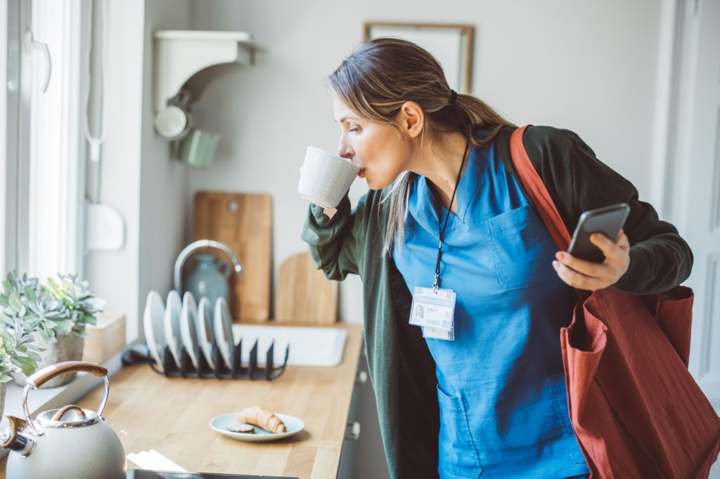 Une femme boit un café à la va-vite au-dessus du plan de travail de sa cuisine. Elle est habillée, prête à partir.