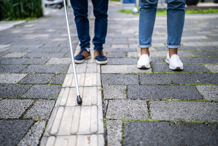 Un aveugle marche avec une canne, dans la rue accompagnée d'une autre personne
