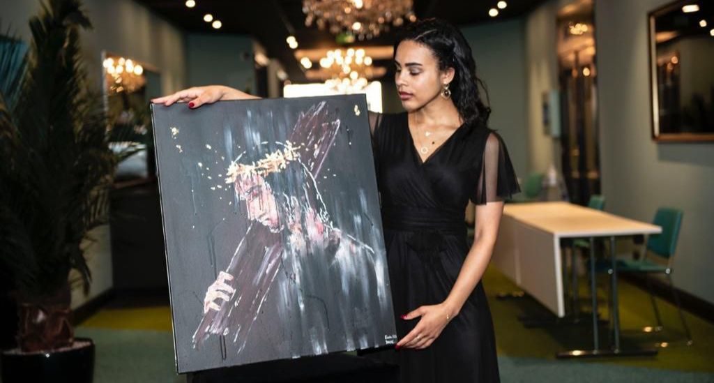 La peintre Haïah tient son tableau intitulé "Esaïe 53", qu'elle présente vers l'objectif. Il représente Jésus portant sa croix sur l'épaule. Le tableau est peint à grand traits et semble inspiré de l'impressionnisme.